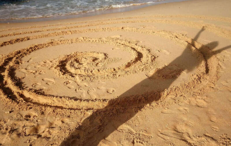 Schatten einer Person mit erhobenen Armen auf einer in den Sand gezeichneten Spirale am Strand, Symbol für Selbstbewusstsein und Persönlichkeitsentwicklung.